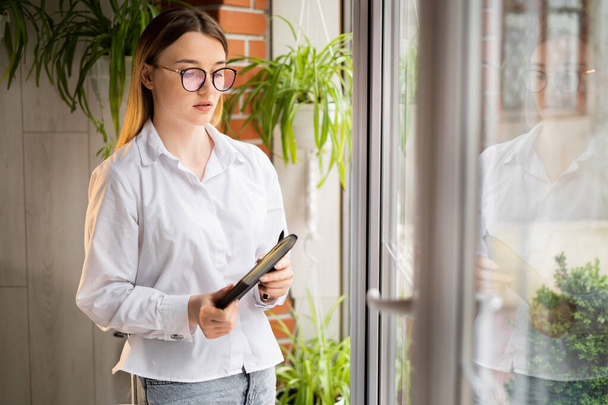 business woman standing in the office by the window with a notebook in her hands. Serious upset secretary with glasses at the panoramic window. Office worker with a paper client base. Work concept. - Фото, зображення