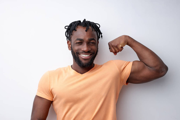 Positive afro american guy in orange t-shirt showing biceps and smiling, isolated - Foto, afbeelding