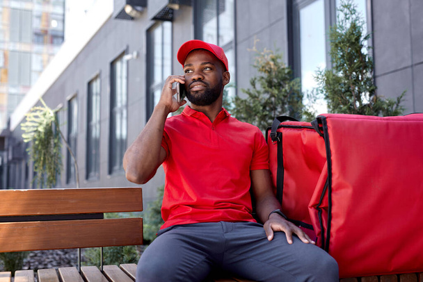 delivery man calling client by mobile phone outdoors. courier carrying bag with food - Zdjęcie, obraz