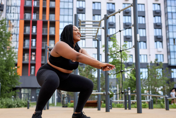 Obese fat african american woman squats in sports ground. Weight losing, obesity - Фото, изображение