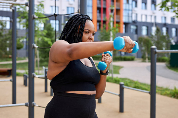 Chubby african woman exercising with dumbbell in public park outdoors, concentrated - Foto, imagen