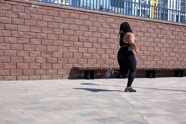 An obese young african female jogging outdoors. Fat beautiful female in black tracksuit - Photo, image