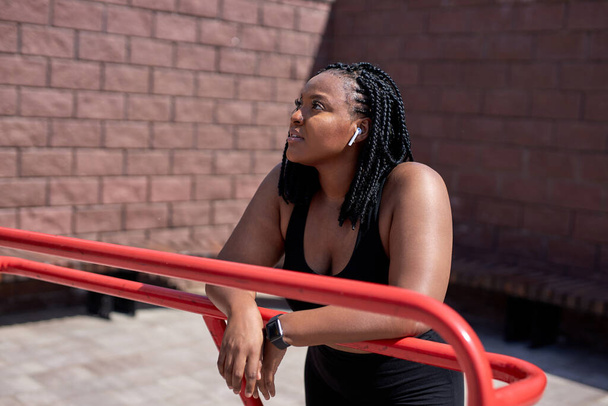 Mujer afroamericana soñadora en ropa deportiva negra descansando, mirando al costado - Foto, Imagen