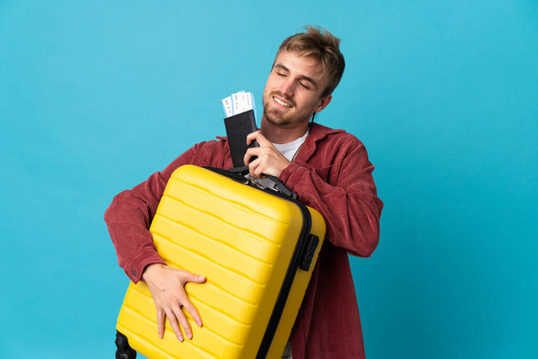 młody przystojny blondynka człowiek odizolowany na niebieskim tle w wakacje z walizką i paszportem - Zdjęcie, obraz