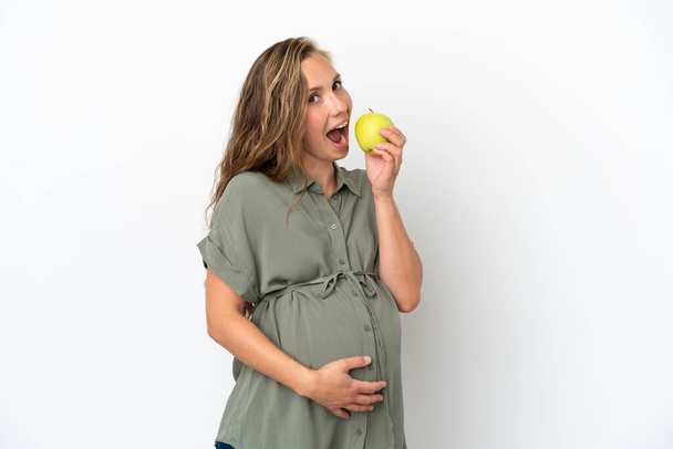 Junge kaukasische Frau isoliert auf weißem Hintergrund schwanger und hält einen Apfel und isst ihn - Foto, Bild