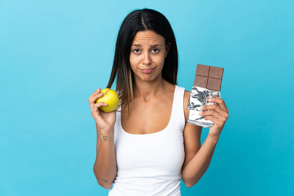 Caucasian girl isolated on blue background taking a chocolate tablet in one hand and an apple in the other - Zdjęcie, obraz