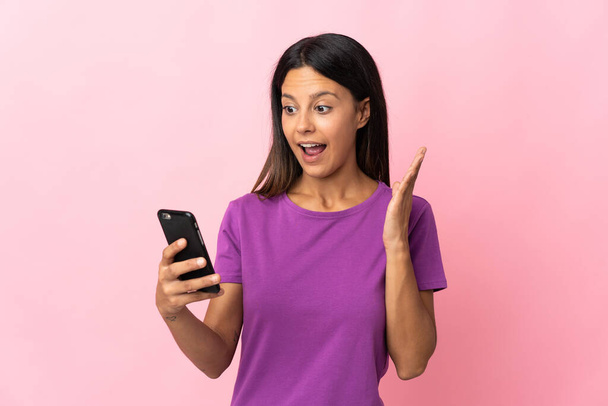 Кавказская девушка выделяется на розовом фоне, глядя в камеру, используя мобильный телефон с удивленным выражением лица - Фото, изображение
