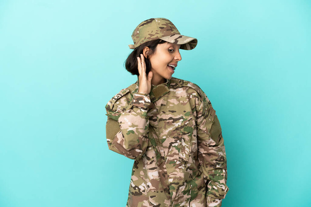 Militar mujer de raza mixta aislada sobre fondo azul escuchando algo poniendo la mano en la oreja - Foto, Imagen