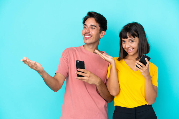 Junges gemischtes Paar hält Handy isoliert auf blauem Hintergrund und streckt die Hände zur Seite, um zum Kommen einzuladen - Foto, Bild