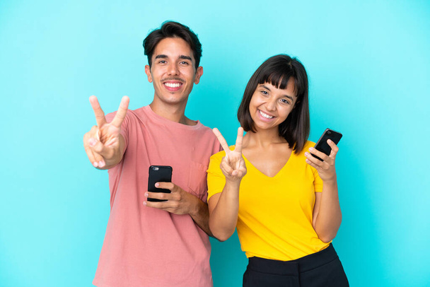 Junges gemischtes Paar hält Handy isoliert auf blauem Hintergrund und lächelt und zeigt Siegeszeichen - Foto, Bild