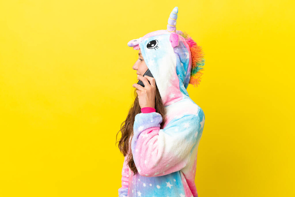 Chica con pijama de unicornio sobre fondo aislado manteniendo una conversación con el teléfono móvil con alguien - Foto, imagen