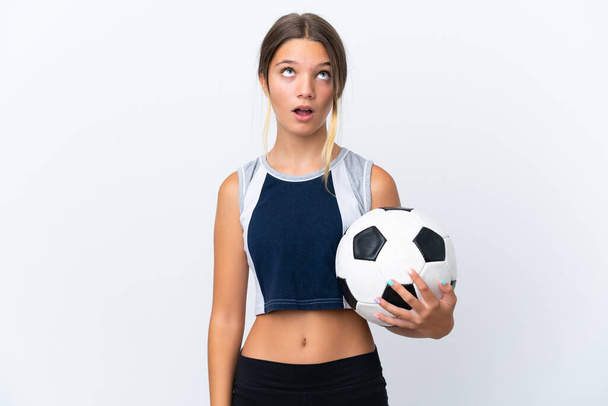 Pequeña chica caucásica jugando fútbol aislado sobre fondo blanco mirando hacia arriba y con expresión sorprendida - Foto, Imagen