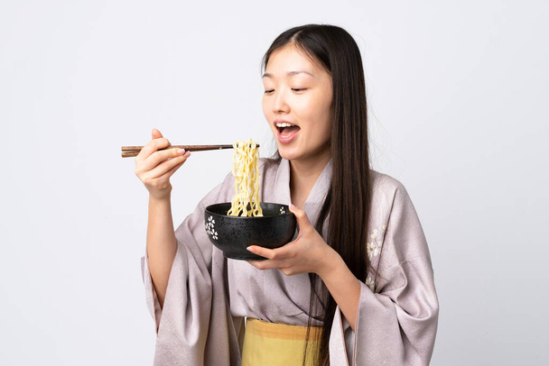 Nuori kiinalainen tyttö yllään kimono yli eristetty valkoinen tausta tilalla kulhoon nuudeleita syömäpuikot ja syö sitä - Valokuva, kuva