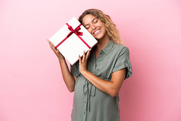 Chica con el pelo rizado aislado sobre fondo rosa embarazada y sosteniendo un regalo - Foto, imagen