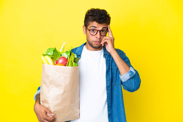 黄色の背景に隔離された食料品の買い物袋を持っている若いブラジル人男性は、アイデアを考えて - 写真・画像