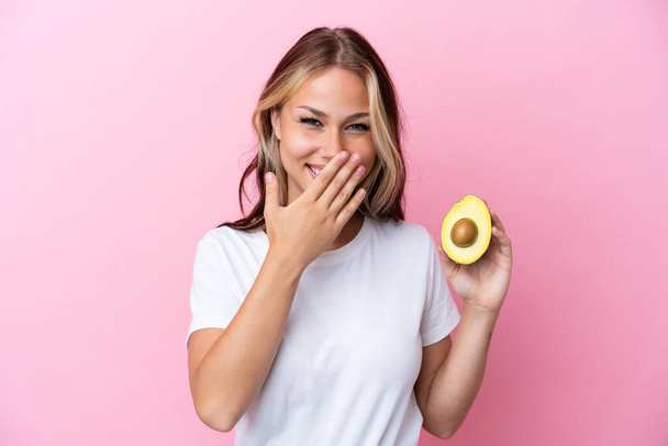 Молода російська жінка, яка тримає авокадо ізольованим на рожевому фоні щасливим і посміхається прикриваючи рот рукою. - Фото, зображення