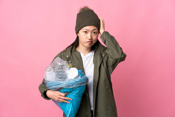 Chica china sosteniendo una bolsa llena de botellas de plástico para reciclar sobre un fondo rosa aislado con una expresión de frustración y falta de comprensión - Foto, imagen