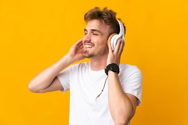 Νέος όμορφος ξανθός άντρας απομονωμένος σε κίτρινο φόντο ακούγοντας μουσική και τραγουδώντας - Φωτογραφία, εικόνα