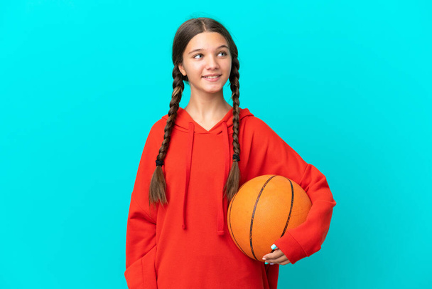 Маленькая кавказская девочка, играющая в баскетбол на голубом фоне, думает об идее, глядя вверх - Фото, изображение