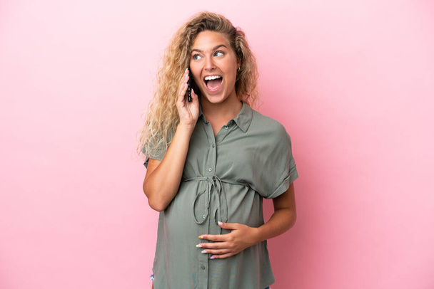 Girl with curly hair isolated on pink background pregnant and using mobile phone with surprised expression - Foto, Imagen