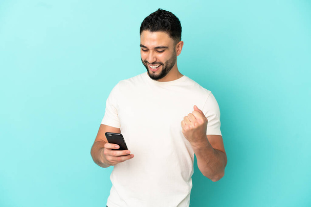 Young arab man isolated on blue background with phone in victory position - Foto, immagini