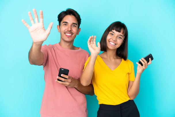 Giovane coppia di razza mista in possesso di telefono cellulare isolato su sfondo blu saluto con mano con espressione felice - Foto, immagini