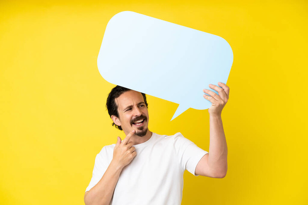 Young caucasian man isolated on yellow background holding an empty speech bubble and pointing it - Photo, Image