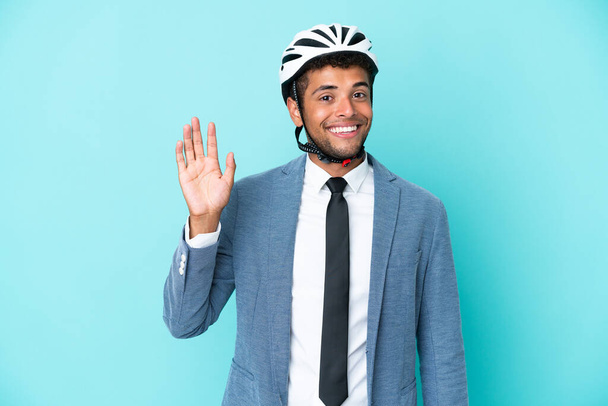 Joven hombre brasileño de negocios con casco de bicicleta aislado sobre fondo azul saludando con la mano con expresión feliz - Foto, imagen