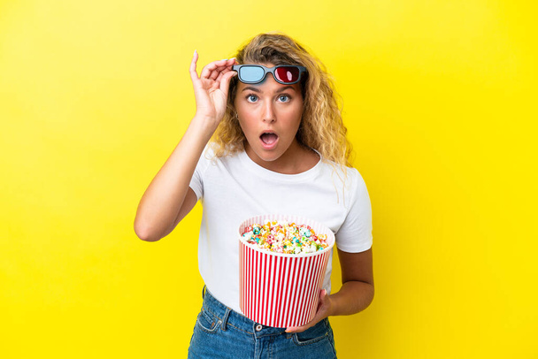 Girl with curly hair isolated on yellow background surprised with 3d glasses and holding a big bucket of popcorns - Foto, Imagen