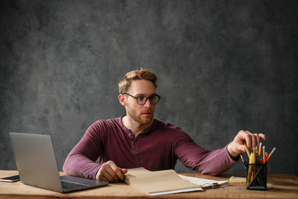 Mies istuu pöydässä papereiden ja kannettavan kanssa valitessaan kirjoituskynää studiossa. - Valokuva, kuva