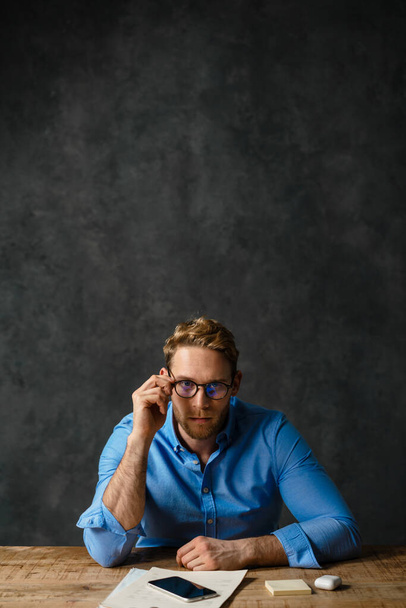 The vertical portrait of a pensive man sitting at the table and adjusting glasses in the studio - Foto, immagini