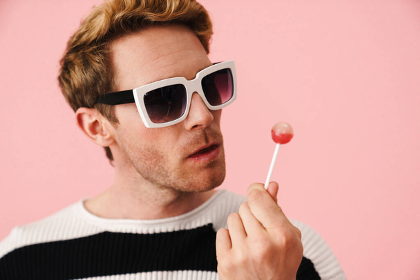 Happy mid aged blonde gay man in sunglasses standing over pink background holding lollipop close up - Фото, зображення