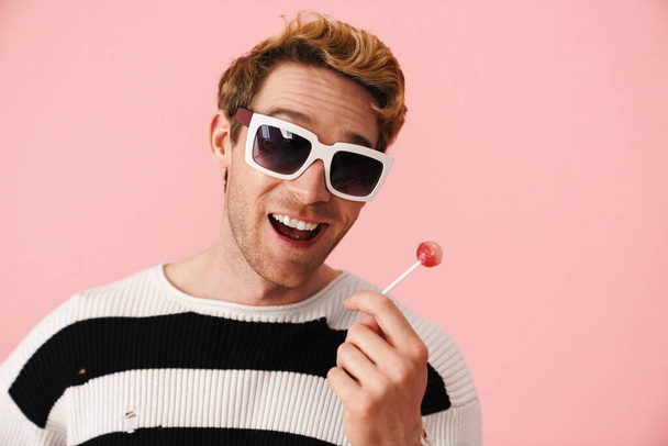 Happy mid aged blonde gay man in sunglasses standing over pink background holding lollipop close up - Foto, immagini