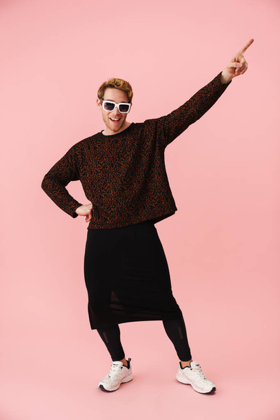 Full length of a happy mid aged blonde gay man in sunglasses standing over pink background posing pointing aside - Foto, afbeelding