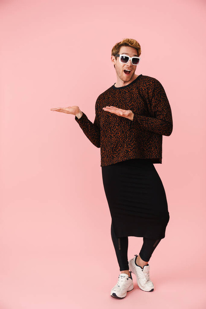 Full length of a happy mid aged blonde gay man in sunglasses standing over pink background pointing aside - Zdjęcie, obraz