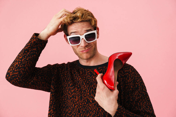 Stylish pensive young white man gay in sunglasses showing high heel red shoes standing over pink background - Fotó, kép