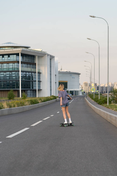 道路上の少女スケートボーダースケート。Tシャツのブロンド女性とジーンズショーツオンロングボード,バックビュー - 写真・画像