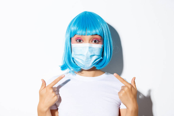 Conceito de celebração do Dia das Bruxas e coronavírus. Close-up de bonito mulher asiática em peruca azul apontando para máscara médica, de pé sobre fundo branco - Foto, Imagem