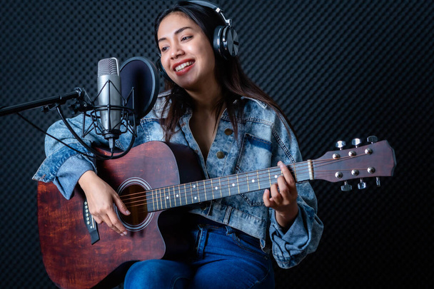 Happy cheerful pretty smiling of portrait a young Asian woman vocalist Wearing Headphones with a Guitar recording a song front of microphone in a professional studio - Фото, зображення
