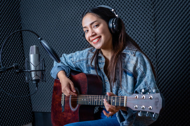 Happy cheerful pretty smiling of portrait a young Asian woman vocalist Wearing Headphones with a Guitar recording a song front of microphone in a professional studio - Fotoğraf, Görsel