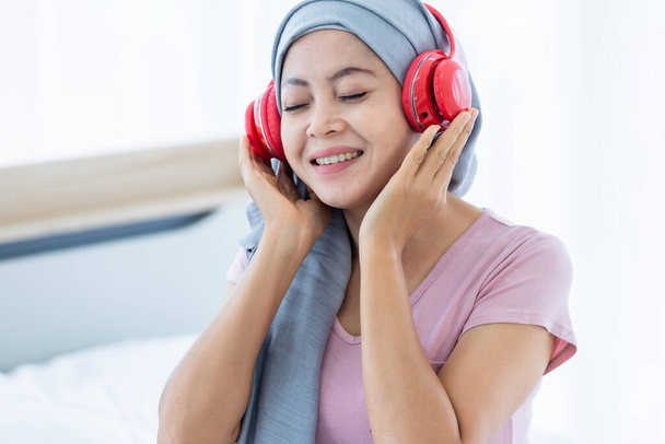 Счастливые азиатские женщины болезнь молочной железы пациент с розовой лентой носить платок в наушниках слушает музыку после лечения химиотерапии сидеть на кровати в спальне в доме - Фото, изображение
