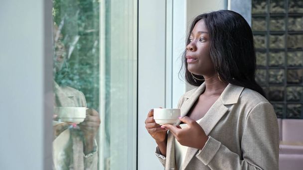 アフリカ系アメリカ人女性がオフィスの窓の近くでコーヒーを飲む - 写真・画像