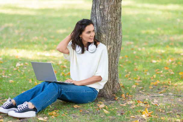 Foto de una mujer de 40 años con portátil sentada en un césped en el parque de la ciudad. - Foto, imagen
