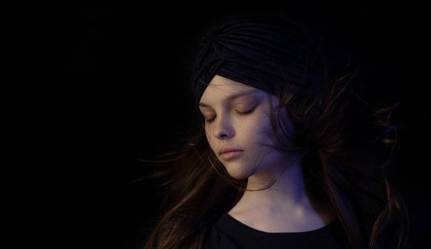 Beautiful little teenager girl with meditative facial expression in multicolor lights. Dark portrait of child with turban and flowing hair shows emotions. Selective focus, low key. - Foto, Imagem