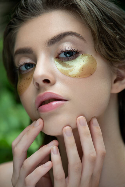 Beautiful young woman applies green  and gold eye mask, closeup. Portrait of beauty model with natural  makeup cares about her skin. Spa, skincare and wellness. Selective focus. - Fotografie, Obrázek