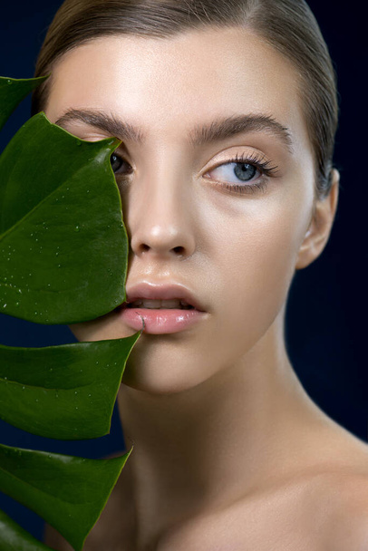 Beautiful young woman's face with perfect skin watching through the tropical green leaves. Beauty model for SPA, wellness and skincare concept.  Close up, selective focus. - 写真・画像