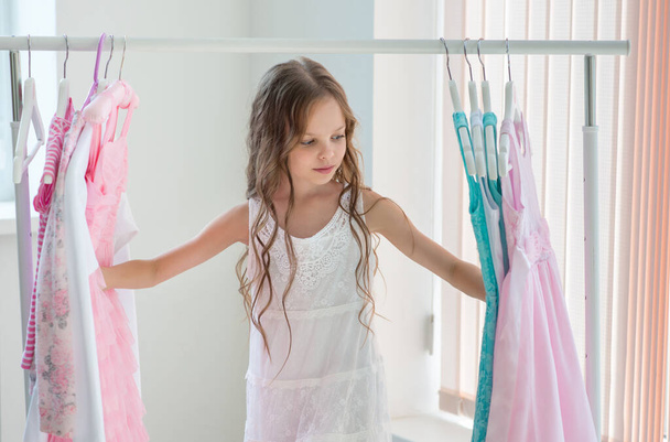 Little child girl choosing her clothes. Kid thinking what to choose to wear in front of many choices of dresses on hangers. Sales, buy and sell concepts. - Photo, Image