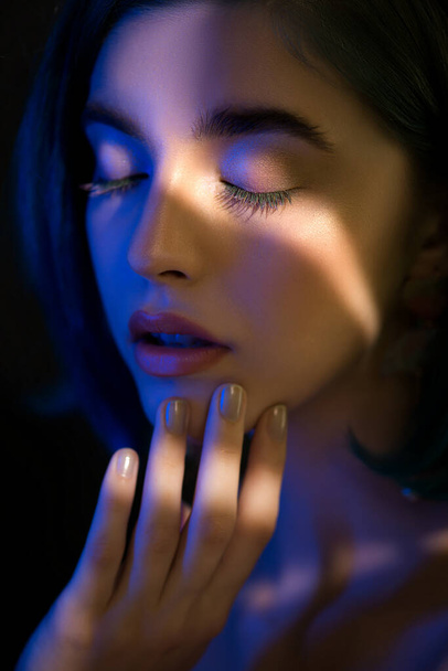 Beautiful young woman with clean perfect skin in neon studio light with light and shadows in her skin. Selective focus. Portrait of beauty model with bright makeup and long eyelashes, SPA and wellness, make-up and skincare concept. - Фото, изображение