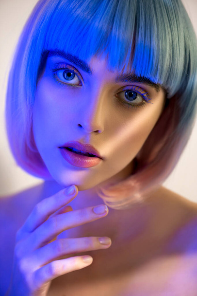 Beautiful young woman in pink and blue wig with clean perfect skin and makeup in neon studio light with light and shadows in her skin. Selective focus. Portrait of beauty model with long eyelashes, SPA and wellness, make-up and skincare concept. - Fotografie, Obrázek