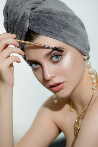 Beautiful young woman with clean perfect skin, pearl earrings and gold jewelry applying makeup. Portrait of beauty model in grey towel with natural classic luxury make up and long eyelashes. - Фото, изображение